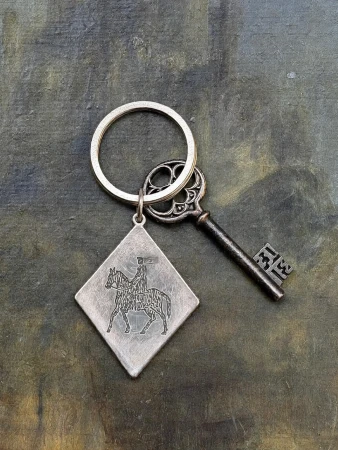 Silver diamond keychain engraved with a knight on horseback attached to an ornate antique key on a textured surface.