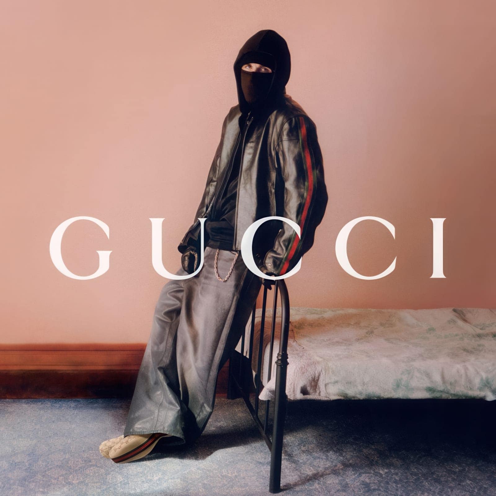 A masked model in a black hooded leather jacket and wide-leg grey trousers leans against a metal bed frame against a peach wall.