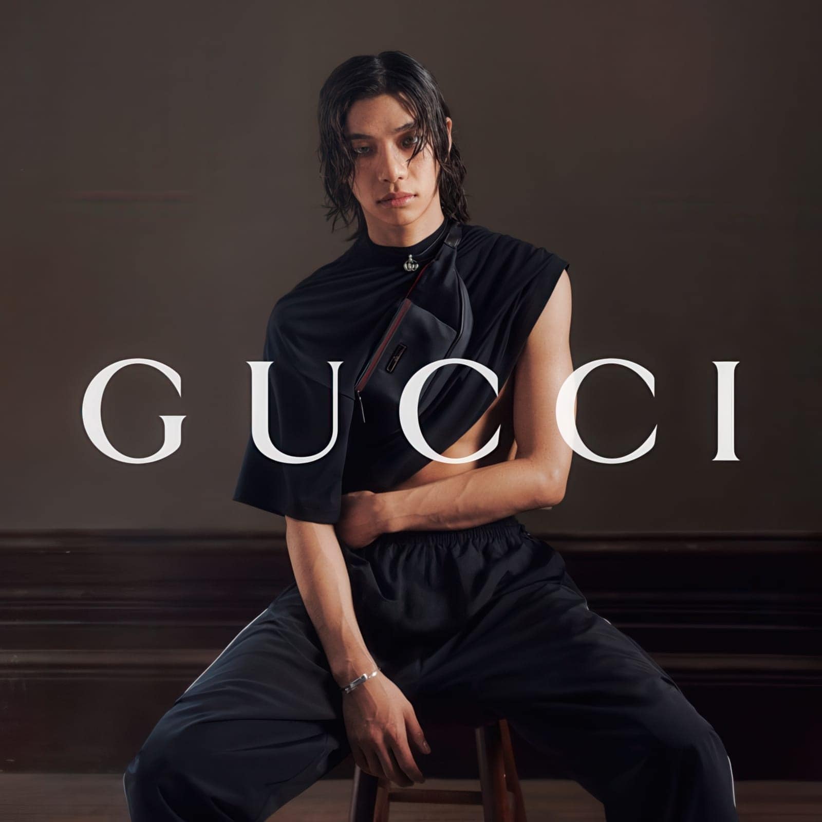 Model in draped black sportswear and crossbody bag seated against a dark backdrop with white Gucci branding.