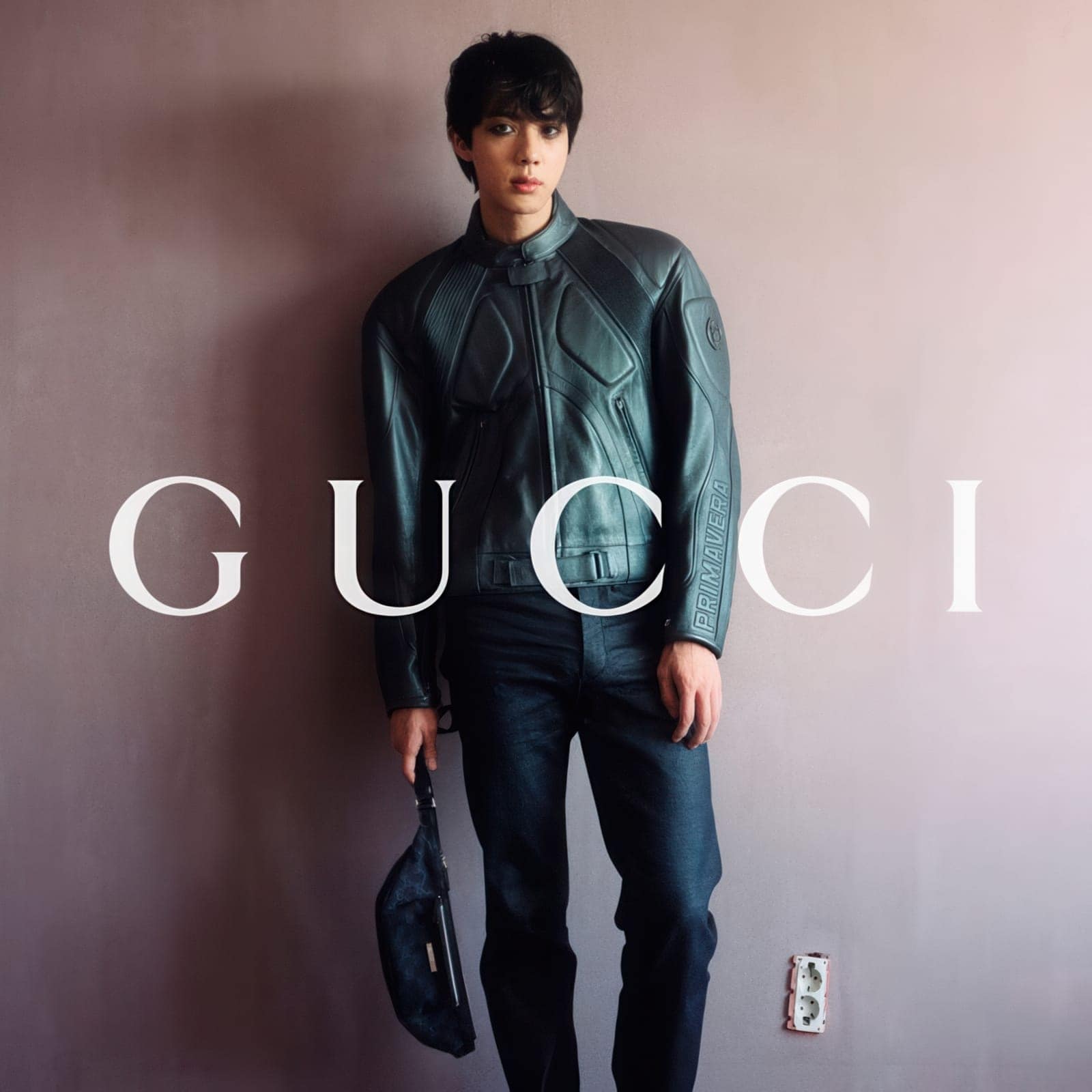 Jin from BTS in a black leather moto jacket and dark jeans holding a handbag for the Gucci The Primavera campaign.