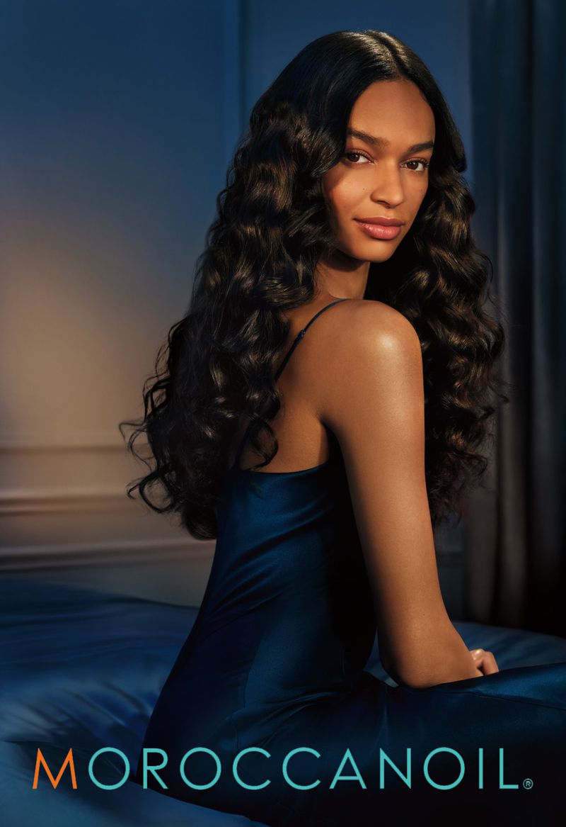 Woman with long, glossy wavy dark hair looking over her shoulder wearing a blue satin slip dress against a blue bedroom backdrop.