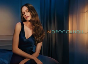 Woman with long wavy brown hair wearing a navy satin dress seated against dark curtains in low evening light.
