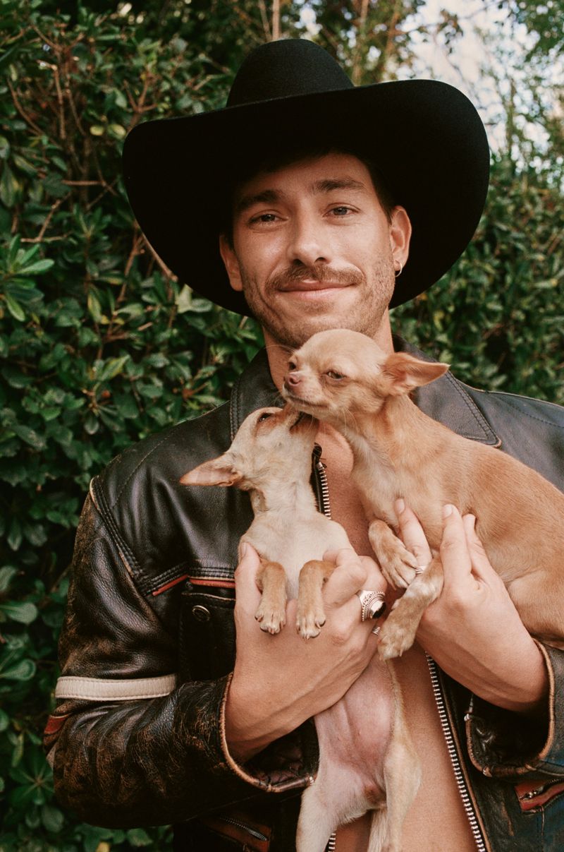 A man in a cowboy hat holds two small dogs in an outdoor setting, wearing a leather jacket and smiling with a mustache and goatee.