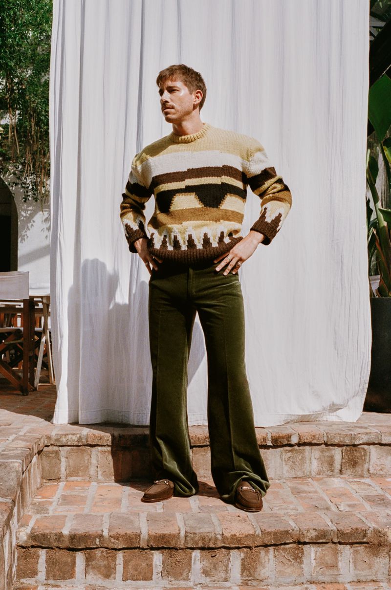 Adult male in olive green corduroy pants and patterned sweater standing on brick steps in front of a white curtain outdoors.