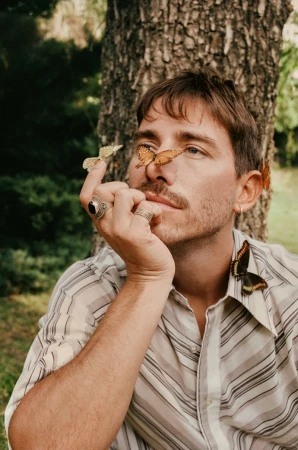 Portrait of a man with butterflies on his face and hand, posing near a tree in an outdoor setting during daylight.