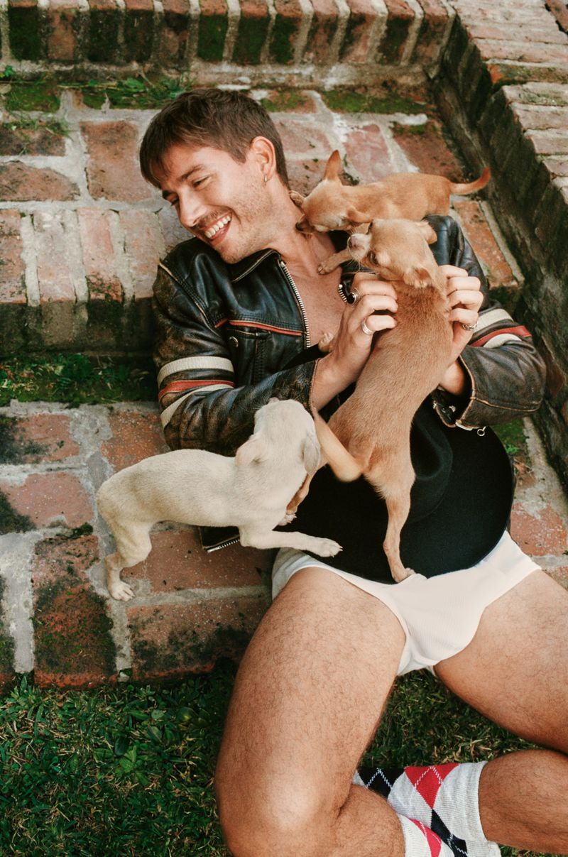 Smiling man lies on brick steps with four small dogs, wearing briefs, argyle socks, and a leather jacket against a garden backdrop.