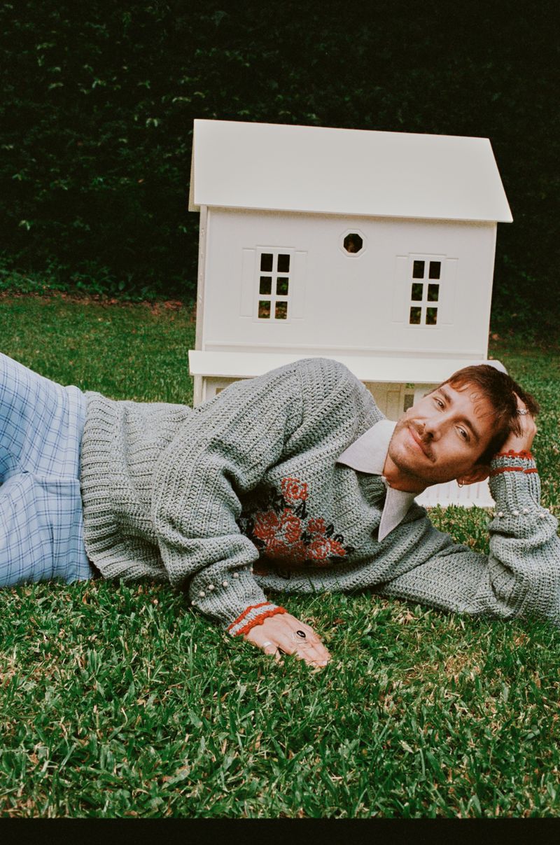 Adult male lying on grass in a sweater with a toy house behind him and greenery in the background under natural lighting.