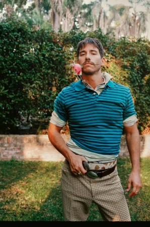 An adult male poses outdoors in a blue and green striped shirt, tan patterned pants, and sunglasses, with a pink rose in his mouth.