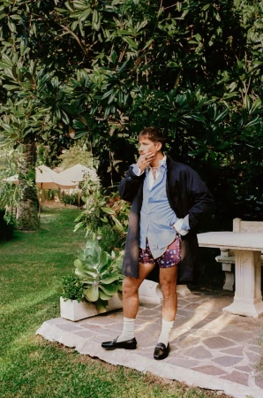 Mature man in outdoor garden setting wearing boxers and a trench coat, accessorized with socks and dress shoes in daylight.
