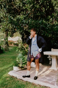 Man standing outdoors in a coat, patterned shorts, white socks, and loafers on a stone patio surrounded by lush garden greenery.
