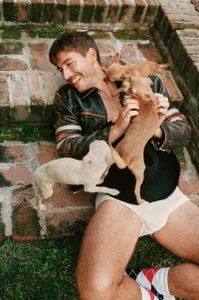 Smiling man in a leather jacket and white underwear playing with three small Chihuahuas on stone steps.