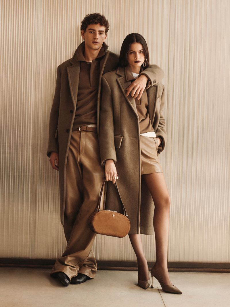 A male and female model pose wearing tailored camel coats, leather bottoms, and pointed heels in front of a fluted wall for Ferrari.