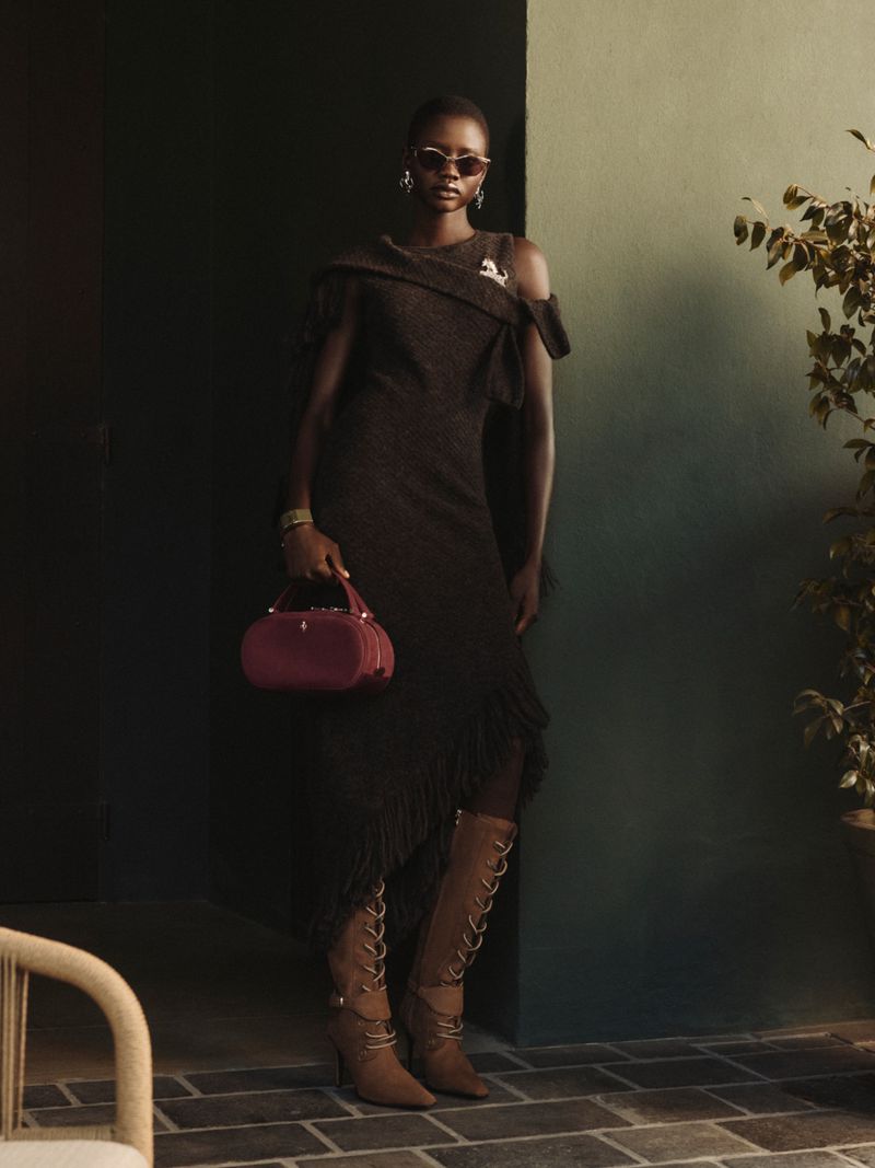 Model posing in a brown off-the-shoulder knit dress, lace-up boots, and sunglasses while holding a burgundy handbag for Ferrari.