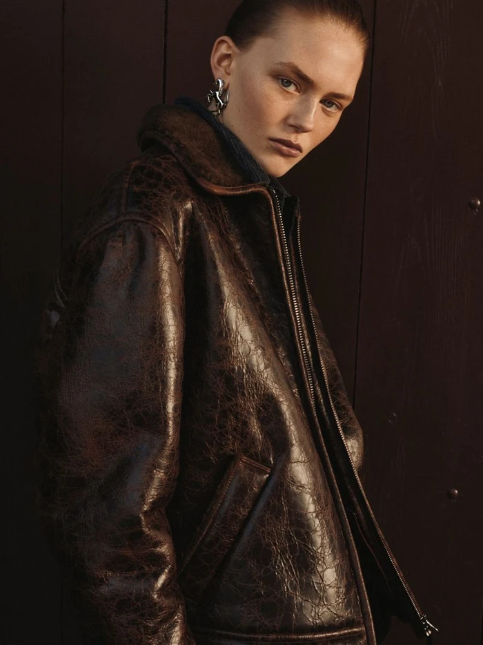Model Agnes Wahlström wearing a dark brown distressed leather bomber jacket and silver sculptural earring against dark wood paneling.