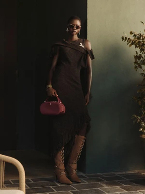 Model wearing a dark textured off-shoulder dress, lace-up knee-high boots, and sunglasses holding a burgundy handbag for Ferrari.