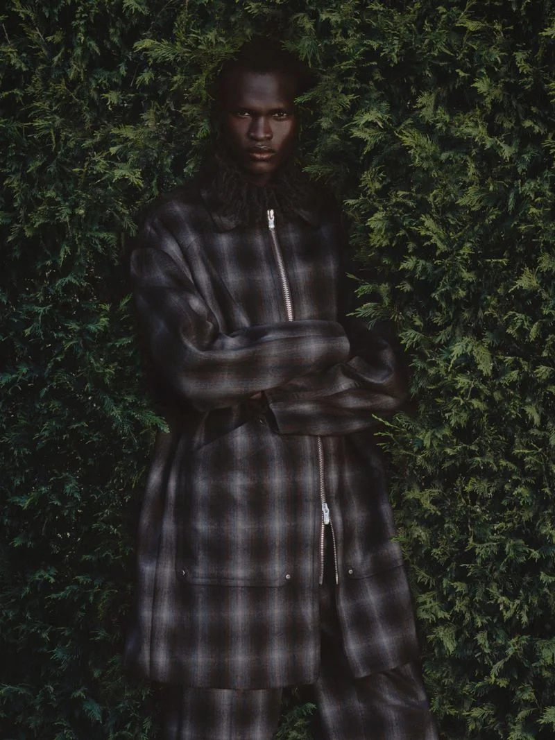 A Black male model wearing a dark plaid zip-up coat poses with arms crossed against a dense green evergreen hedge.