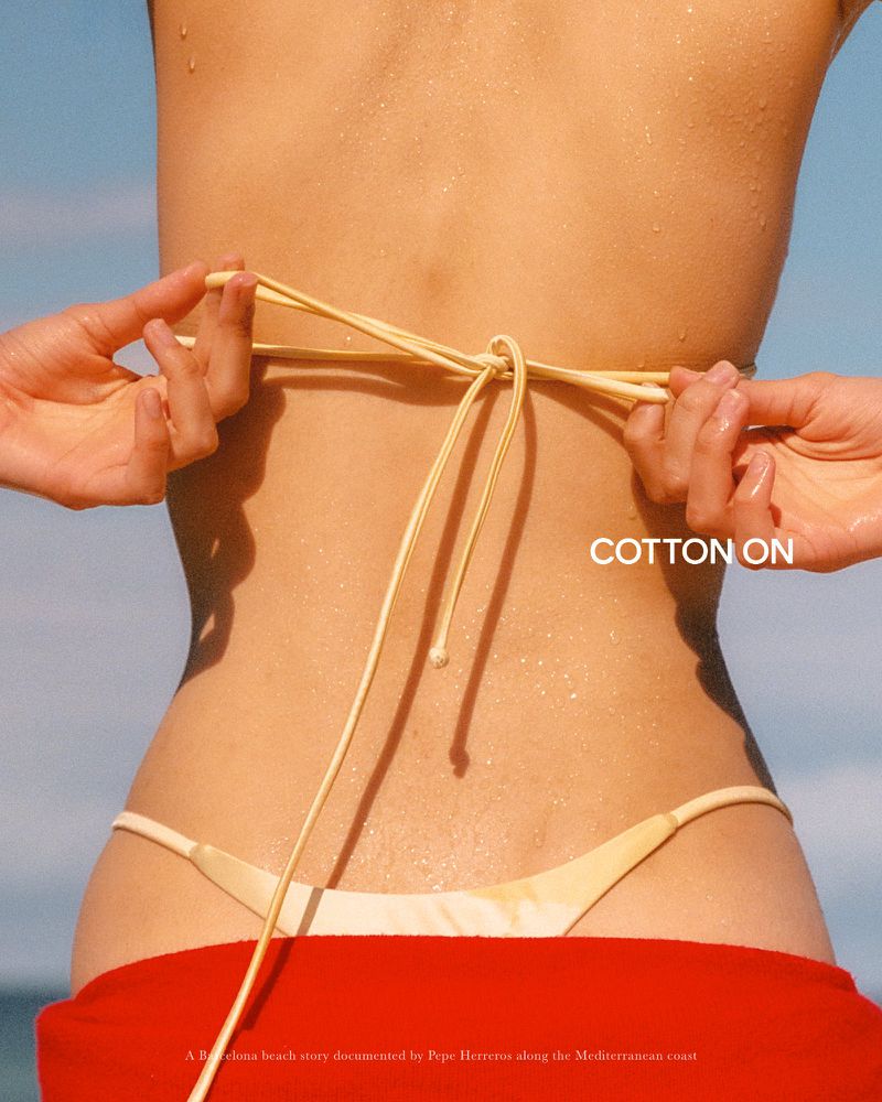 Close-up from behind of a woman adjusting the back ties of a cream string bikini with a bright red wrap around her waist in sunlight.