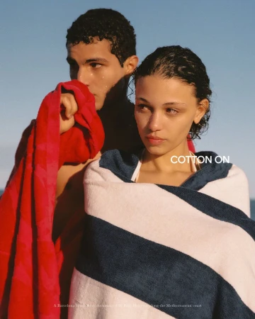 A woman wrapped in a navy and white striped towel next to a man holding a red beach towel under direct warm sunlight.