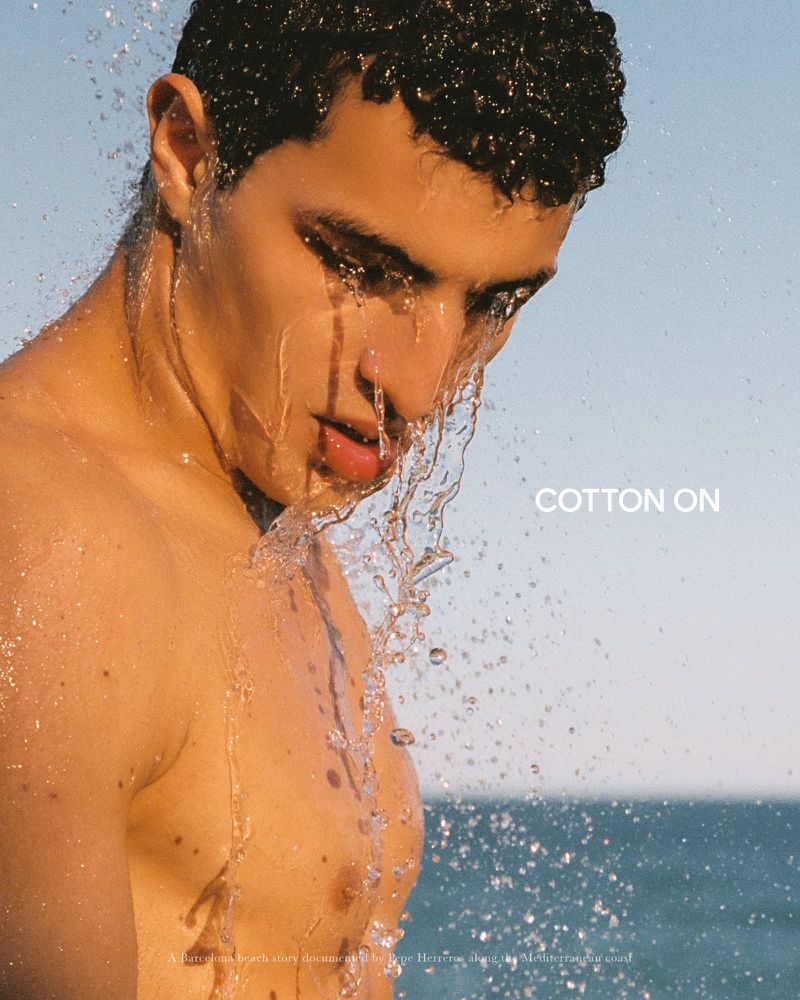 Close-up of a young shirtless man with water splashing across his face in front of an open ocean on a sunny day.