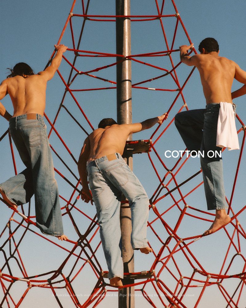 Three shirtless young men wearing blue denim jeans climb a red rope playground structure under a clear blue sky.