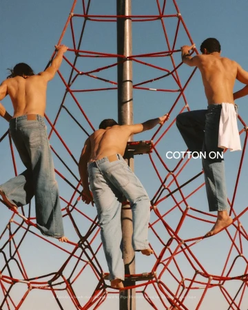 Three shirtless young men wearing blue denim jeans climb a red rope playground structure under a clear blue sky.