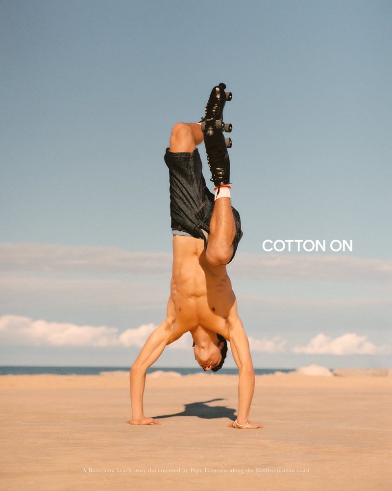 Shirtless man performing a handstand wearing black roller skates and denim shorts on a sunny beach.