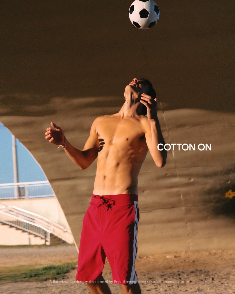 Shirtless man in red board shorts looking up at a soccer ball in mid-air outdoors beneath a concrete bridge structure.