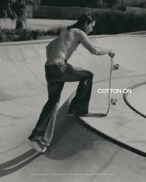 Black and white photograph of a shirtless man in flared denim jeans holding a skateboard on the edge of a concrete skatepark bowl.