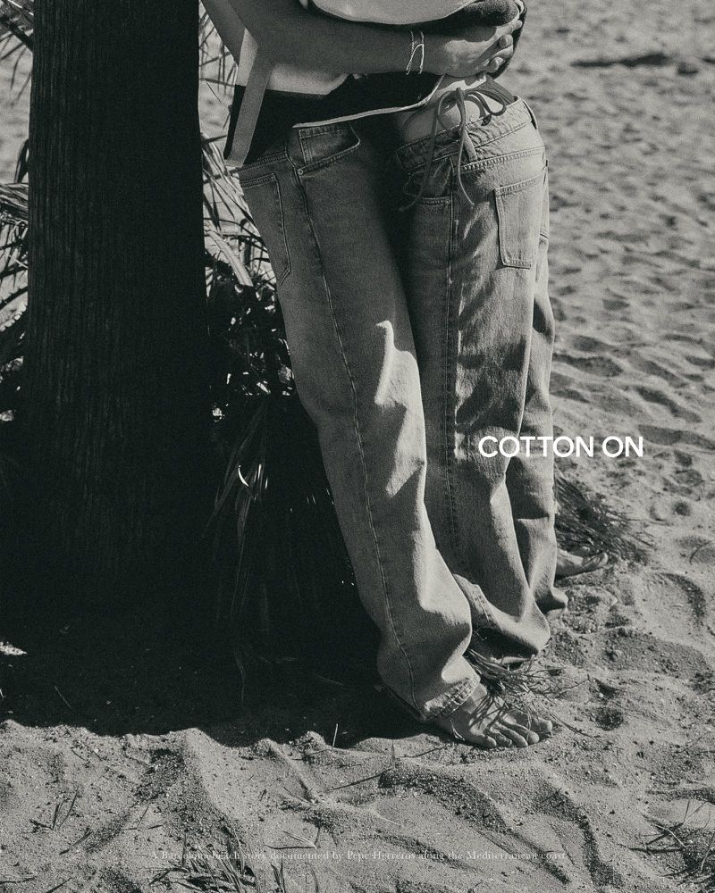 A black and white shot of two people in denim jeans standing barefoot on a sandy beach next to a tree.