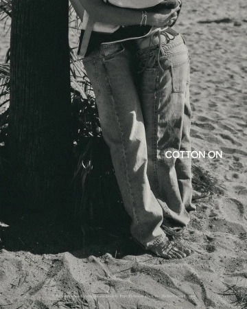 A black and white shot of two people in denim jeans standing barefoot on a sandy beach next to a tree.