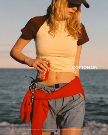Model wearing a raglan tee and blue shorts with a red sweater tied around her waist holding a soda can at the beach.