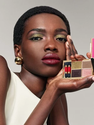 Black female model with shimmering green eyeshadow holding an open Carolina Herrera gold eyeshadow palette against a grey studio backdrop.