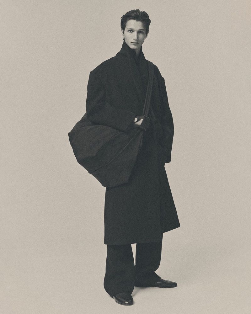 Full-length studio portrait of a male model wearing an oversized long black coat, wide trousers, and carrying a large black crossbody shoulder bag.