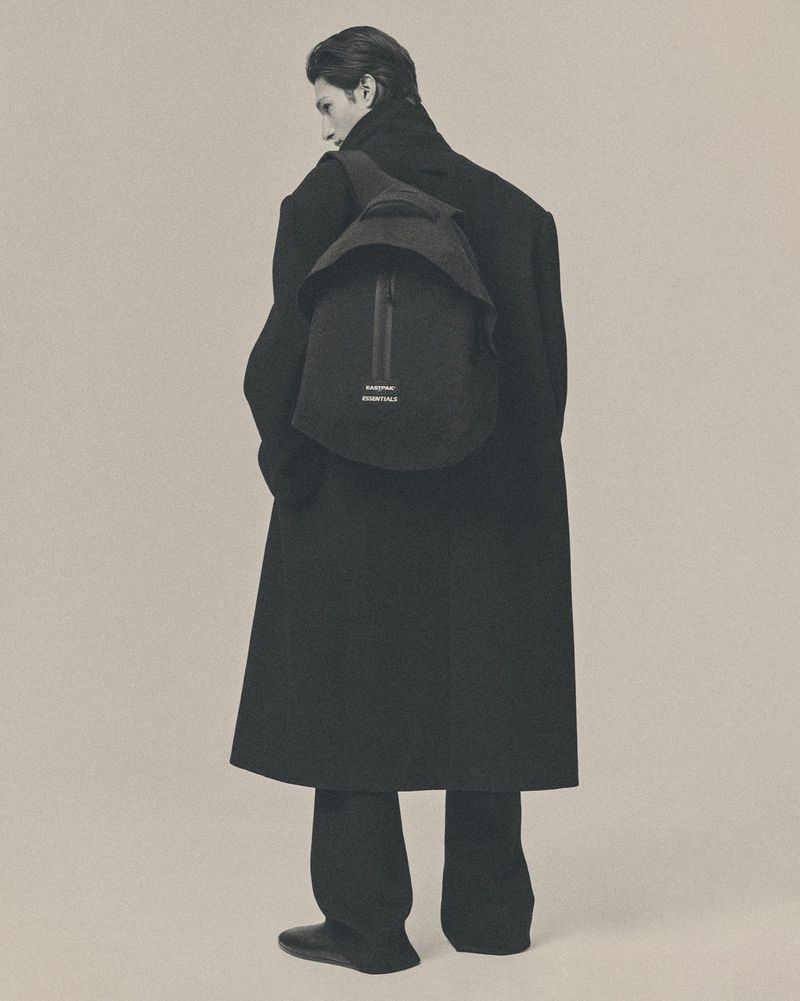 A man turned away wearing an oversized black tailored overcoat, black trousers, and a black Eastpak Essentials backpack.