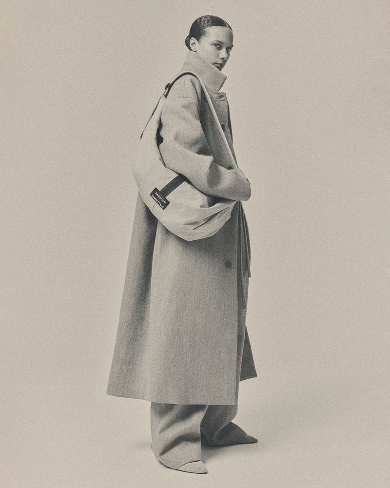 Model in a high-collar oversized beige coat and matching wide-leg trousers wearing a large crossbody bag against a neutral backdrop.