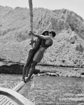 A man in swim trunks and a woman in a bikini embrace on the bow of a sailboat against a rocky coastal background in black and white.
