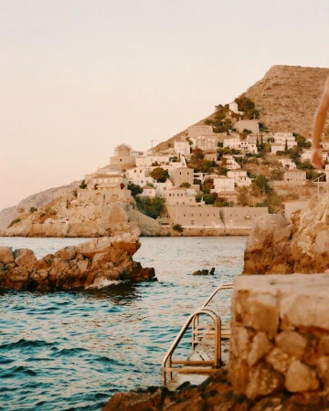 Coastal view of a rocky swimming ledge with a metal ladder leading into deep blue water facing a hillside stone village across the bay.