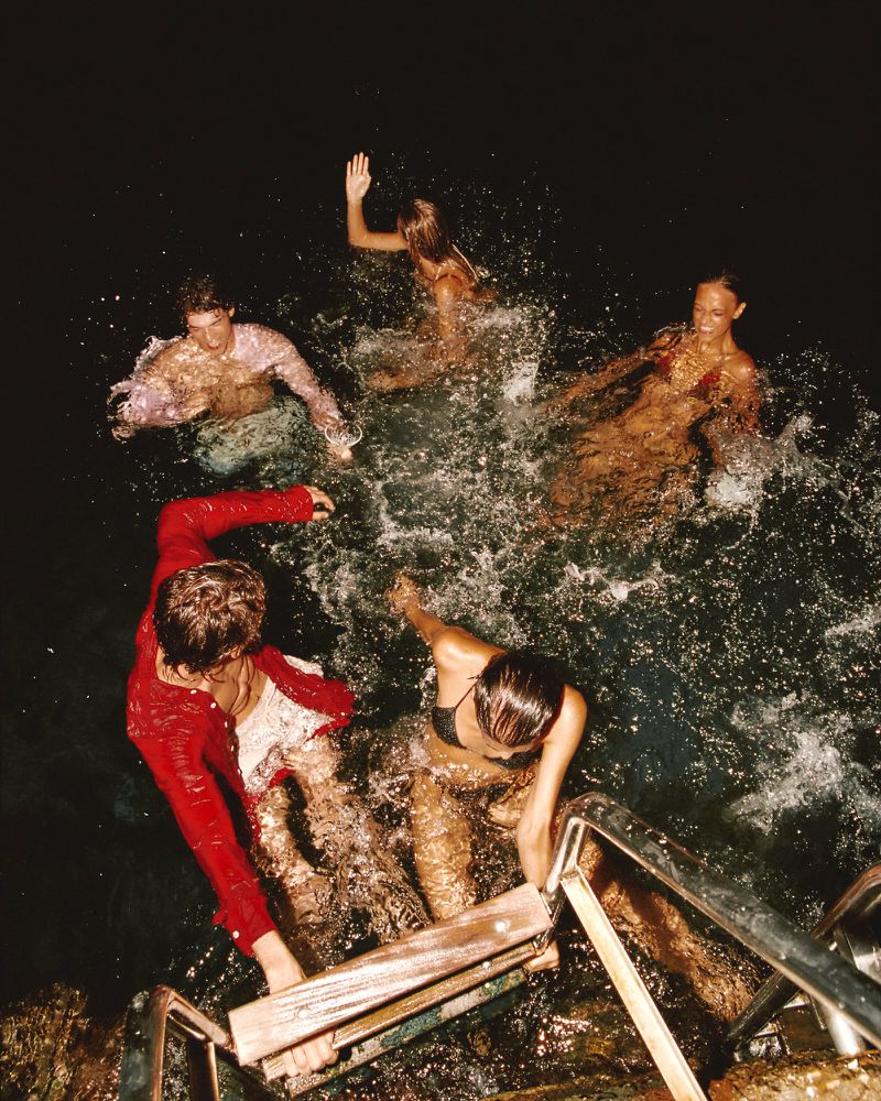 Group of young people swimming in the dark ocean at night with a man in a red shirt climbing a boat ladder under direct flash lighting.