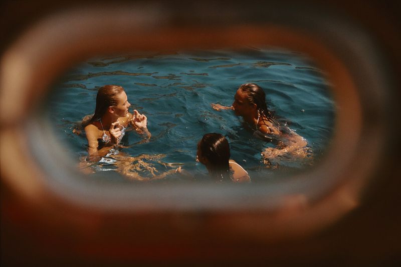 Three swimmers in deep blue open water viewed through a warm, rounded boat porthole.