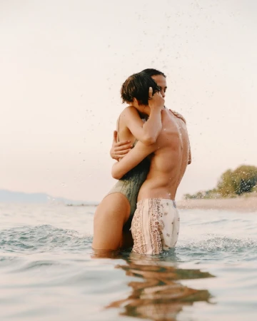 A man and woman embrace waist-deep in ocean water with droplets splashing around them in soft natural light.