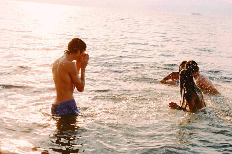 Young man in blue swim trunks standing in ocean water with friends swimming nearby under golden sunlight.