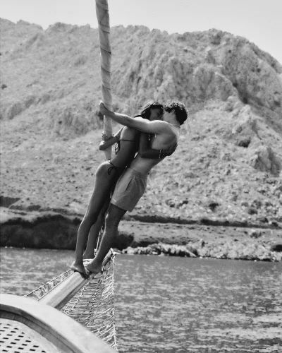 A man and woman embracing on the bowsprit of a boat against a backdrop of rocky hills and open water in black and white.