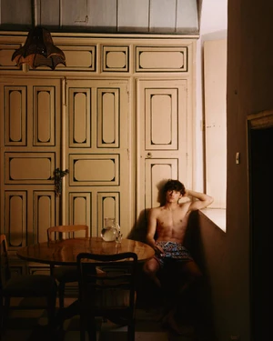 Gonzalo Allgayer sits shirtless in patterned swim shorts by a sunlit interior window beside a wooden table and paneled cupboard.