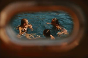 Three swimmers in deep blue open water viewed through a warm, rounded boat porthole.