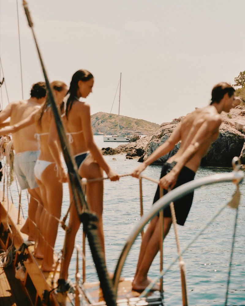 Group of models in swimwear standing on the wooden deck rail of a boat overlooking a rocky coastal cove.