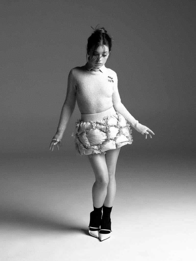 Full body portrait of a young adult woman in studio lighting, wearing a sweater and skirt, styled with heels and socks in black and white.