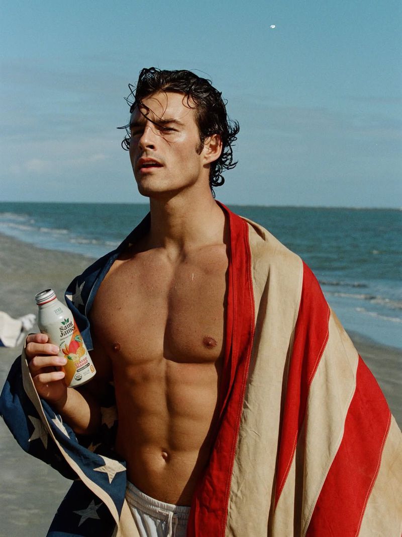 Shirtless man with wet hair draped in an American flag towel, holding a bottle of Saint James Iced Tea on a sunny beach.