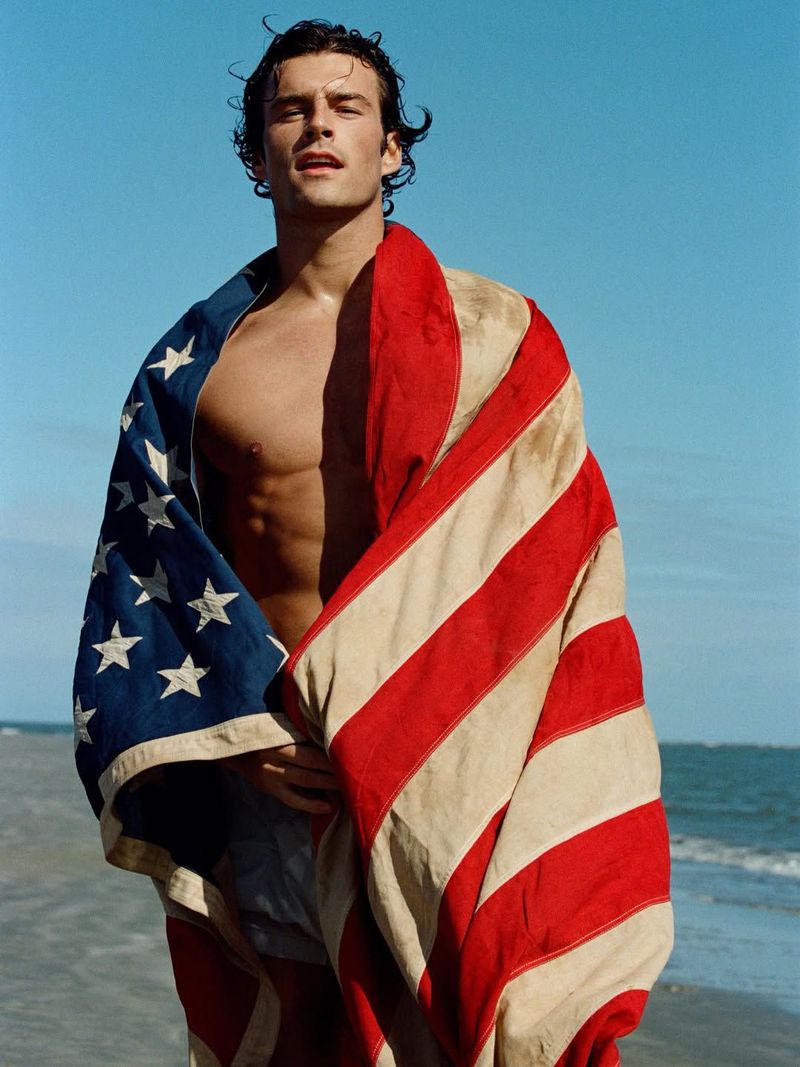 Shirtless man wrapped in an American flag on a sunny sandy beach with blue ocean and sky.