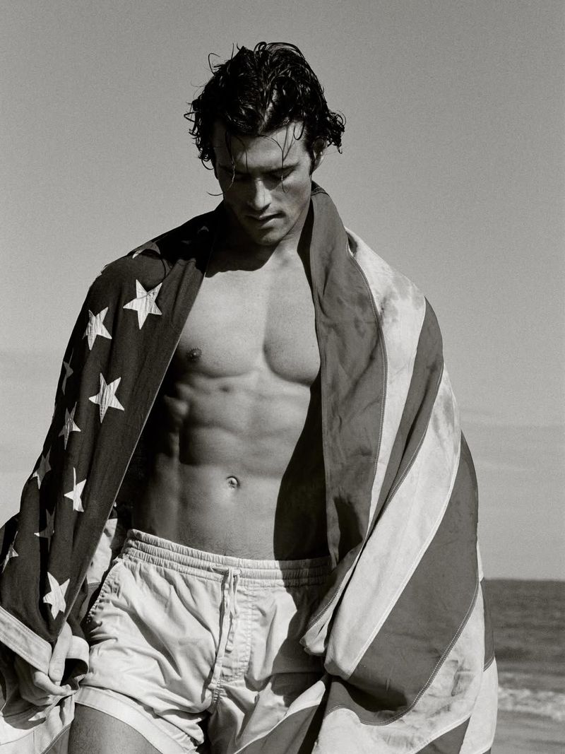 Black and white portrait of model Aiden Stonehouse draped in an American flag wearing swim shorts on a sunny beach.