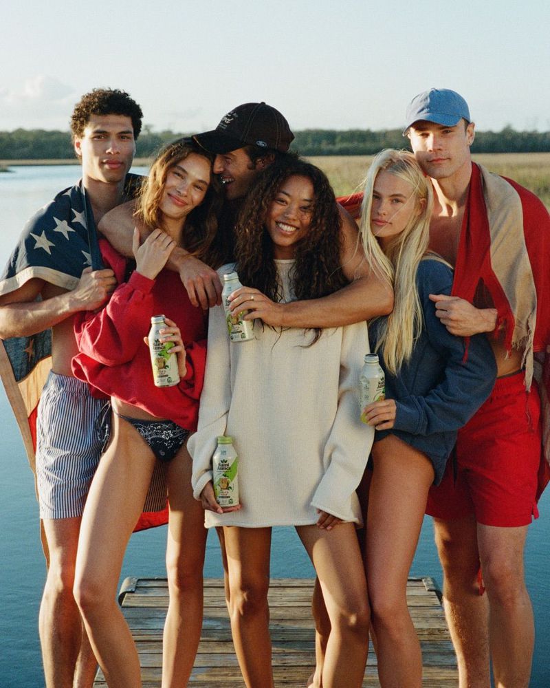 Group of five young adults smiling and posing on a wooden dock by the water holding iced tea bottles.
